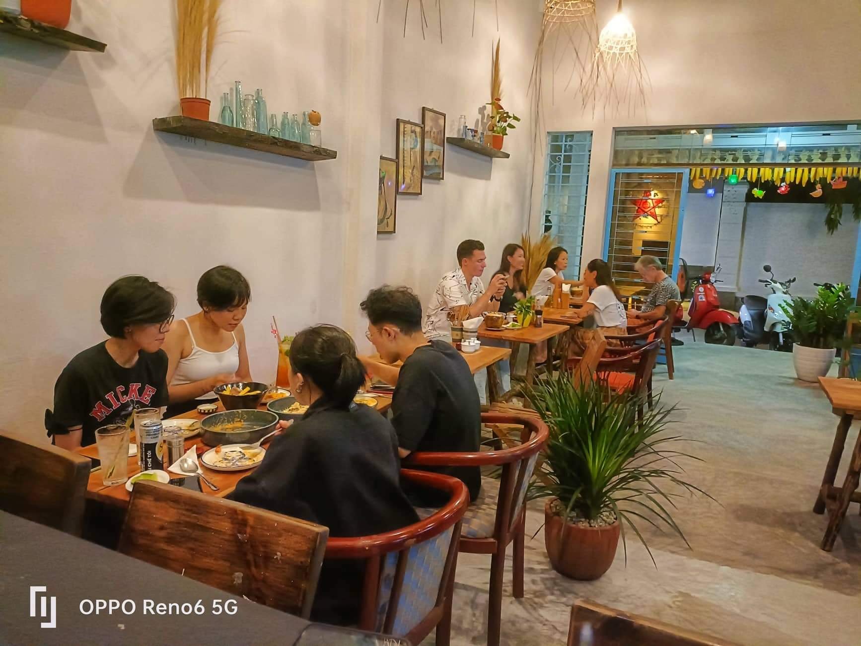 The image depicts an indoor dining area where several groups of people are seated at wooden tables enjoying a meal together. The decor includes shelve