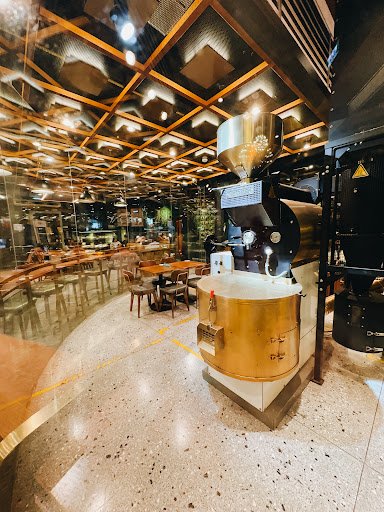 The image depicts the interior of a modern café or coffee shop in Thao Dien, Saigon. It features a prominent coffee roasting machine with a golden bas