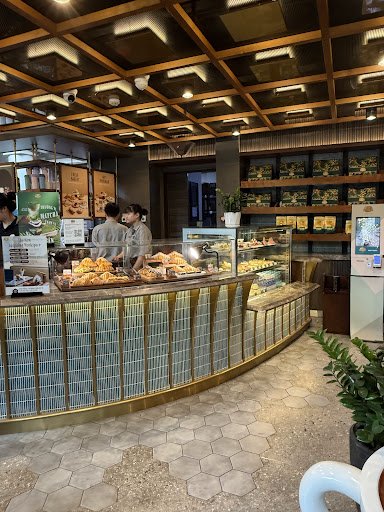 The image shows an interior view of a modern bakery or café counter area where staff are attending to customers and displaying various baked goods on