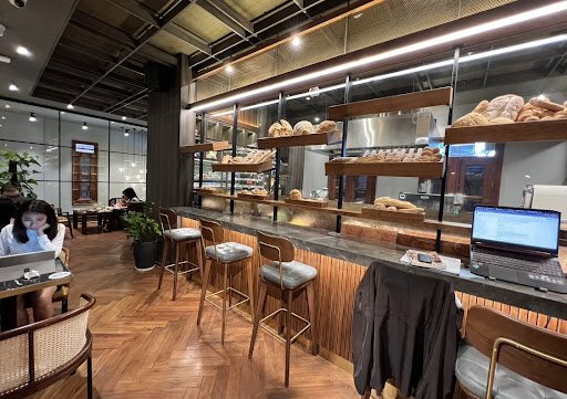 The image depicts an interior view of a modern café or bakery that features wooden flooring and shelving displaying various baked goods such as breads