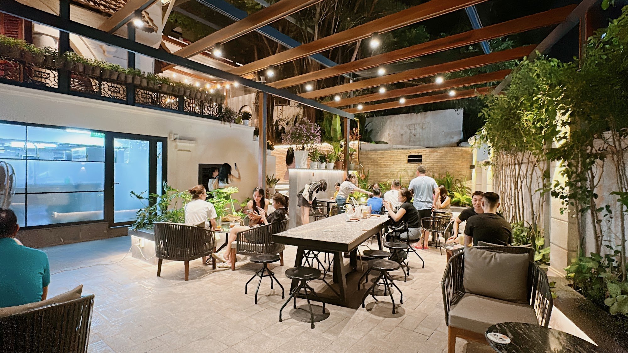 The image depicts an outdoor dining area of a venue in Thao Dien, Saigon, featuring a covered patio with wooden beams and string lights. Multiple tabl