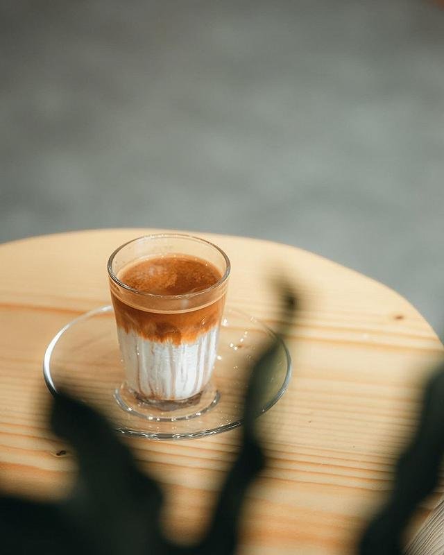 A layered drink in a glass on a wooden surface, possibly coffee-based given the color and texture of the layers.