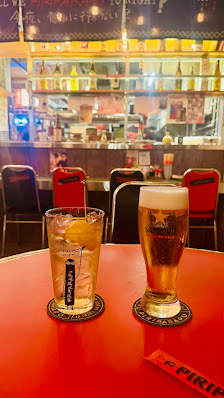 Two drinks on a red table: a tall glass of beer with foam and a short glass with ice and a lemon slice. Background shows a bar with shelves of bottles