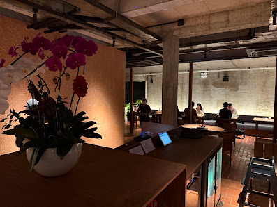 A modern interior space with exposed concrete ceilings and industrial elements. Features wooden tables, chairs, a vase with pink orchids on a table, a
