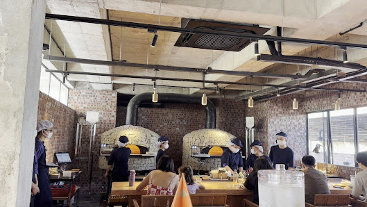 The image depicts an interior view of a restaurant or pizzeria where patrons are seated at tables while staff members work in the background near larg