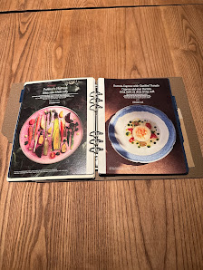The image shows a spiral-bound menu placed on a wooden surface displaying two pages of food items with images and descriptions.