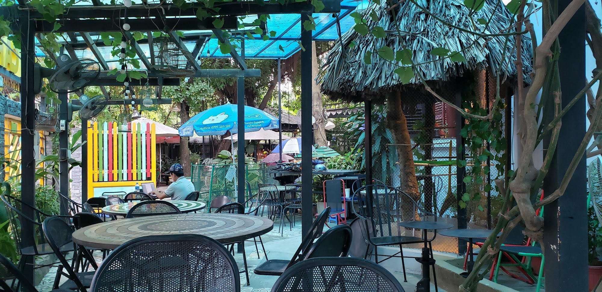 The image depicts an outdoor seating area of a restaurant or café in Thao Dien, Saigon. The setting is vibrant and colorful with various tables and ch