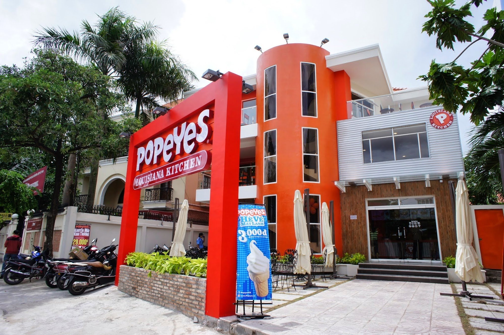 The image depicts the exterior of a Popeyes Louisiana Kitchen restaurant located in Thao Dien, Saigon. A prominent red sign with "Popeyes Louisiana Ki
