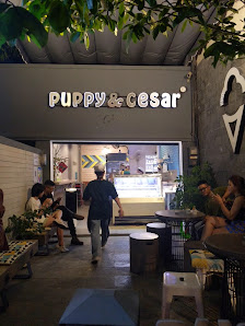 The image shows an exterior view of a cafe named 'Puppy & Cesar' with patrons sitting outside on chairs and tables, enjoying their time.