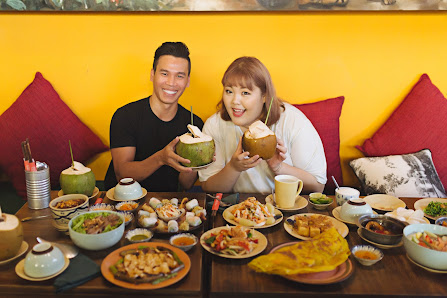 A couple seated at a wooden table filled with various Vietnamese dishes, including coconuts, spring rolls, fried items, and other plates. The backgrou