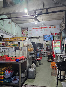 The image depicts the interior of a bustling food shop in Thao Dien, Saigon. It features a narrow space with tiled flooring, various kitchen equipment