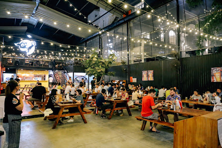 The image depicts a bustling indoor venue in Thao Dien, Saigon, featuring a spacious industrial-style interior with high ceilings, exposed beams, and 