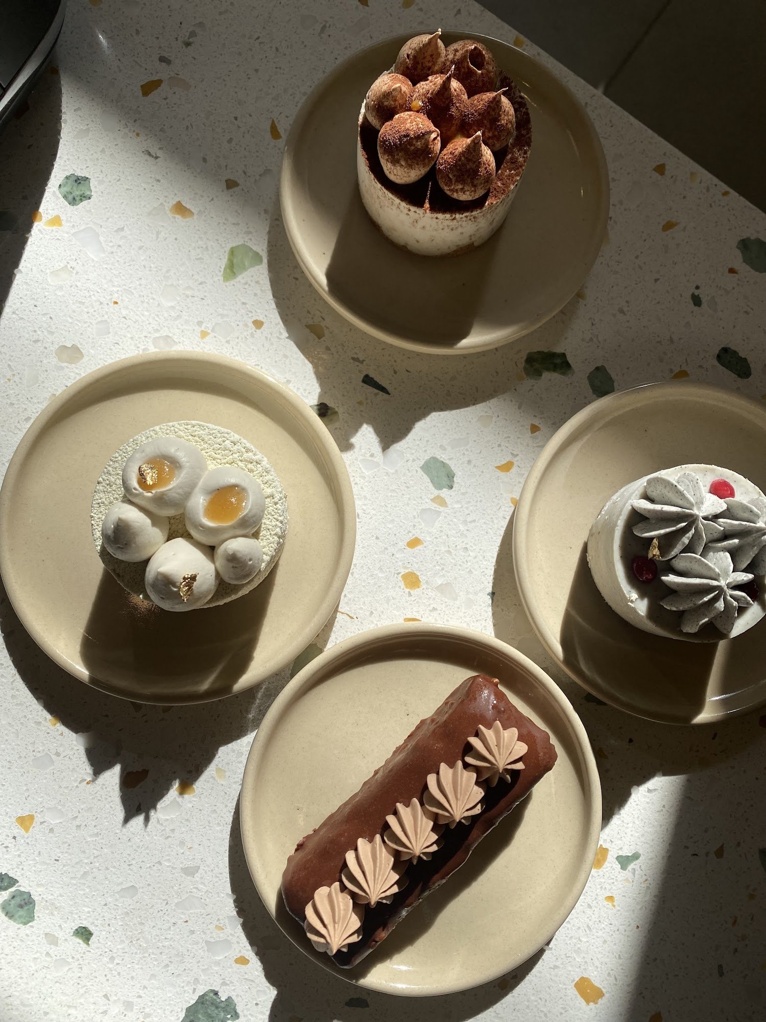 A selection of desserts including a chocolate-covered pastry with leaf-shaped decorations, a cake topped with what appears to be coffee beans and cocoa powder, a dessert cup garnished with white cream flowers and red berries, a plate featuring two egg custard buns on top of a powdery substance, and another dessert resembling a flower made from whipped cream.