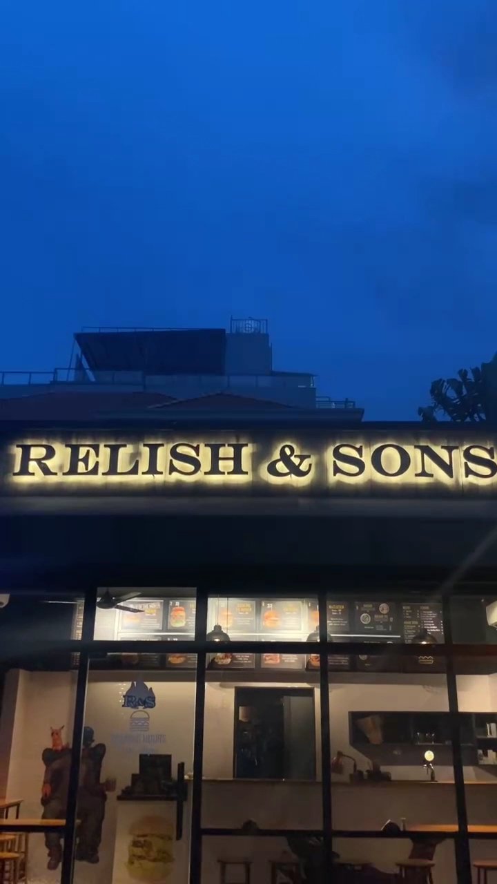 The image shows an exterior view of a restaurant named 'RELISH & SONS' during the evening with illuminated signage above its entrance.