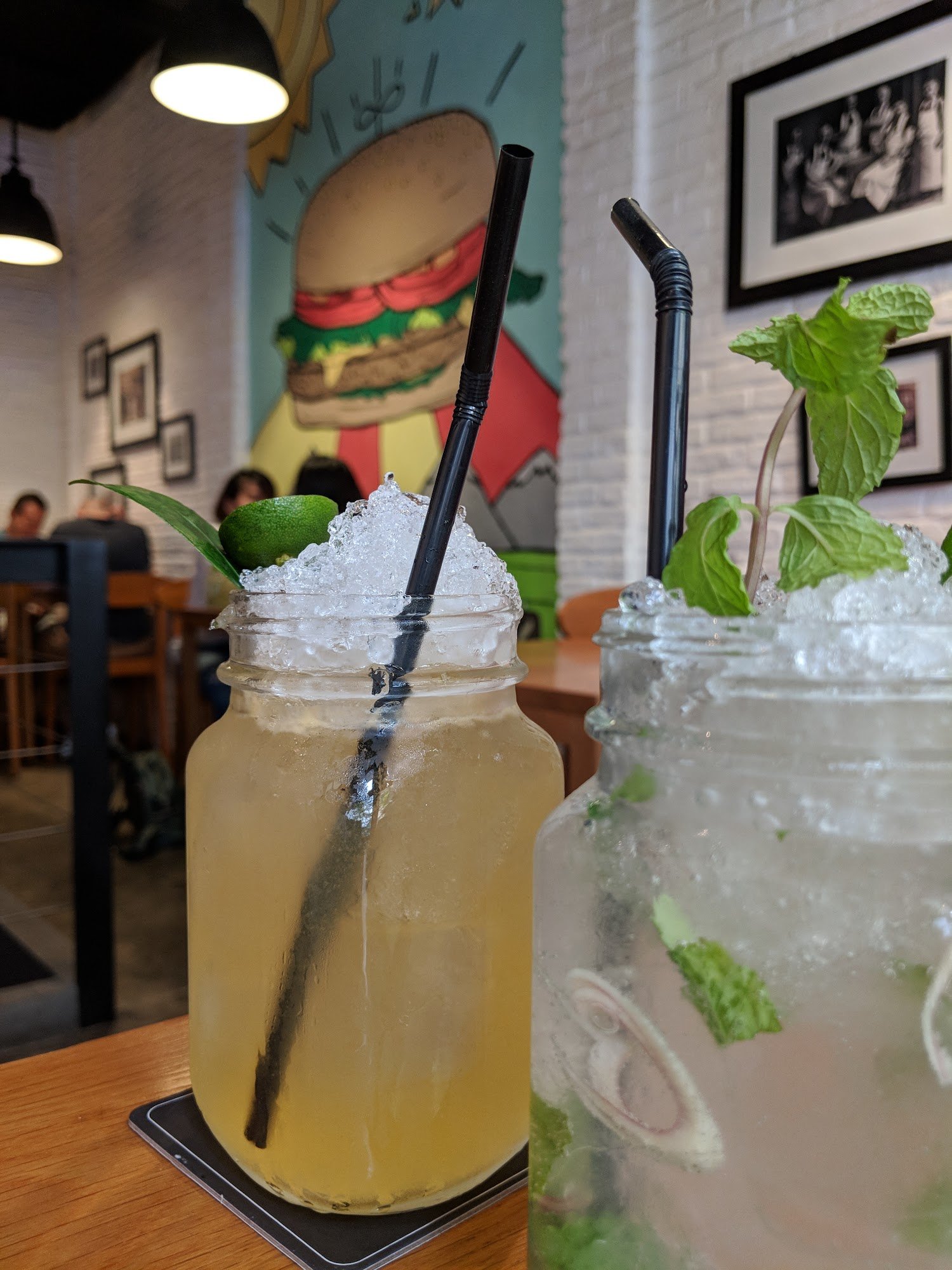 The image shows a close-up of two beverages on a table in an indoor setting with modern decor and artwork depicting food items, including a burger.