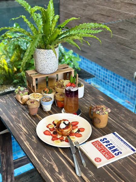 A wooden outdoor table by a pool with various items: a plate of pancakes topped with strawberries, bananas, and cream; multiple small dessert cups (in
