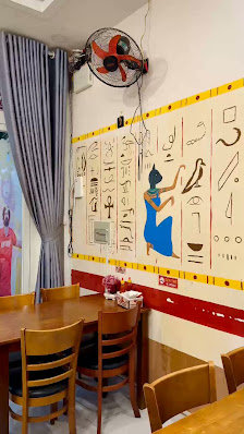 The image shows an interior of a restaurant or cafe featuring wooden tables and chairs arranged for dining. The walls are decorated with colorful Egyp
