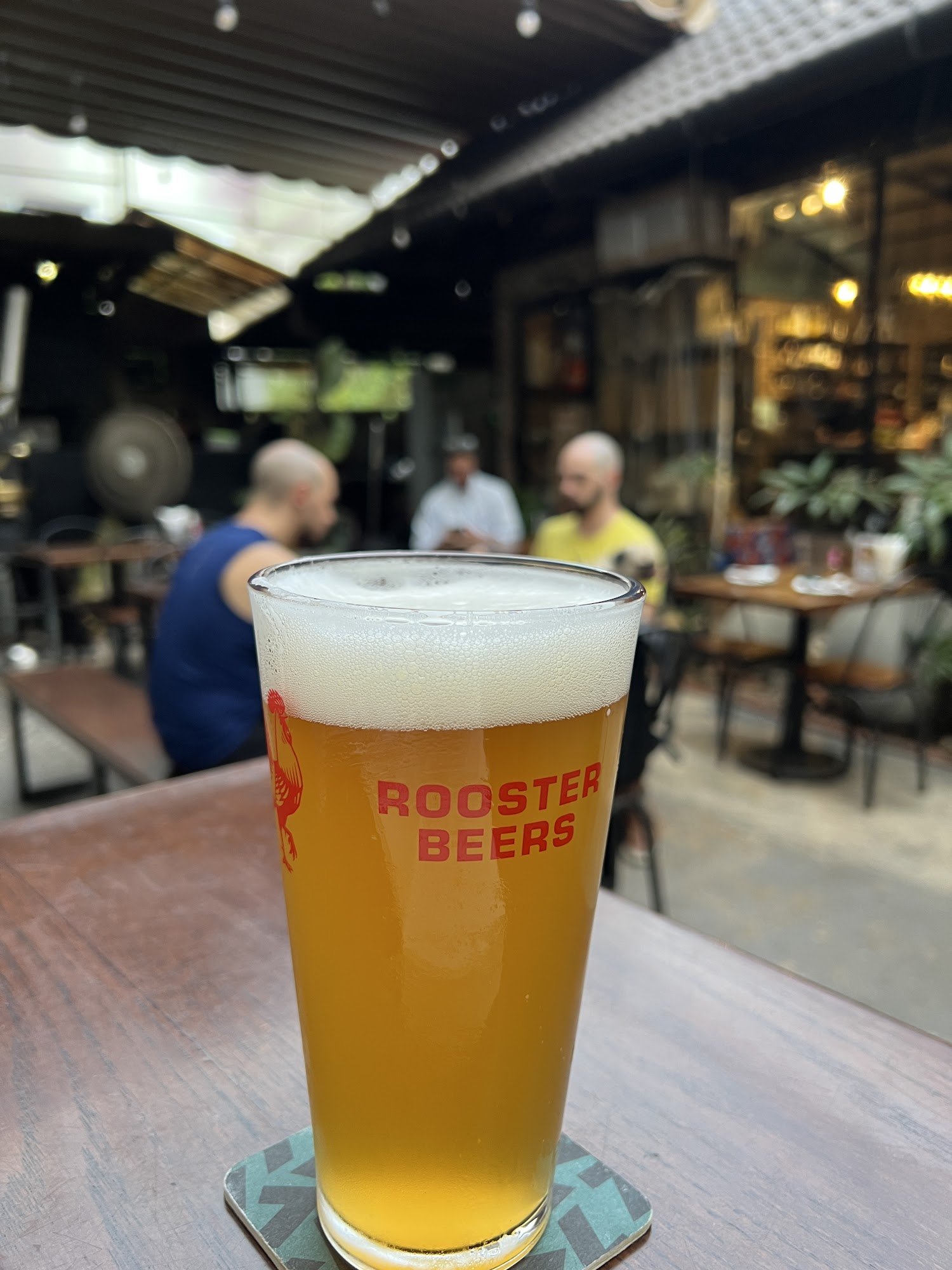 A close-up of a beer glass branded 'Rooster Beers' on a table at an outdoor seating area in Thao Dien, Saigon.
