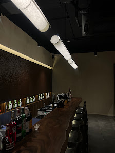 The image depicts an interior view of a modern bar or lounge area featuring a long wooden counter with various bottles and glasses on display, under w