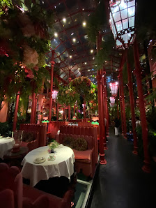 An indoor venue with red upholstered chairs, white tablecloths on round tables, lush green plants, red pillars, a glass ceiling with hanging lights, a