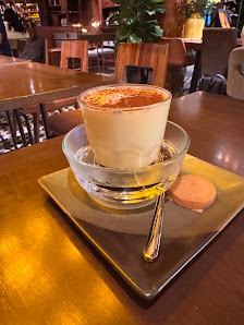 A cup of coffee or latte art sits on a saucer in an indoor setting with wooden furniture and warm lighting, suggesting a cozy café ambiance.