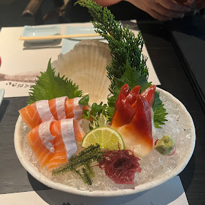 A plated dish of sashimi featuring various types such as salmon and tuna served on a bed of ice with garnishes like lime, wasabi, pickled ginger, seaw