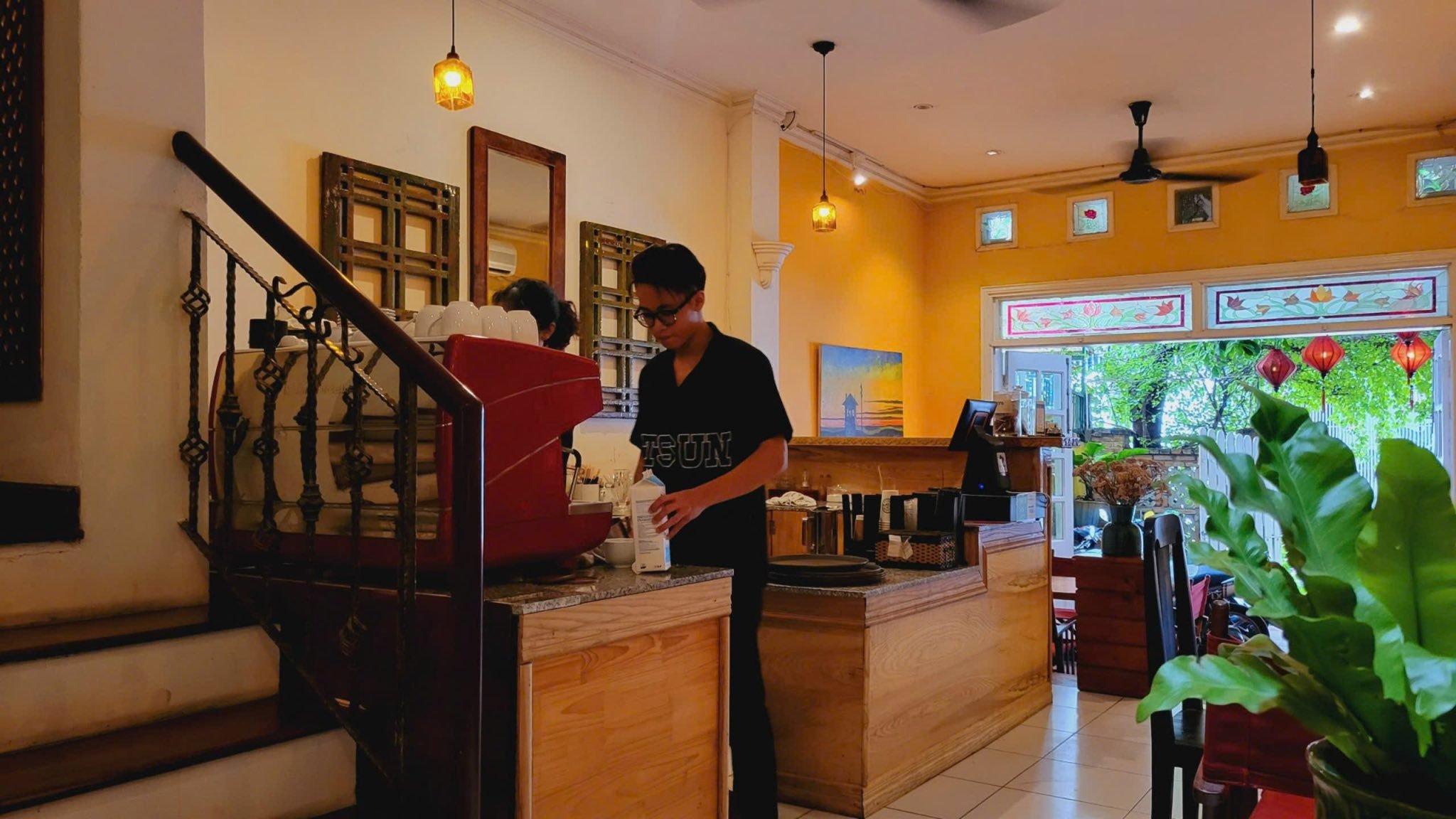 The image depicts an interior view of a cozy café or restaurant setting. A person is seen working behind the counter with various items such as cups a