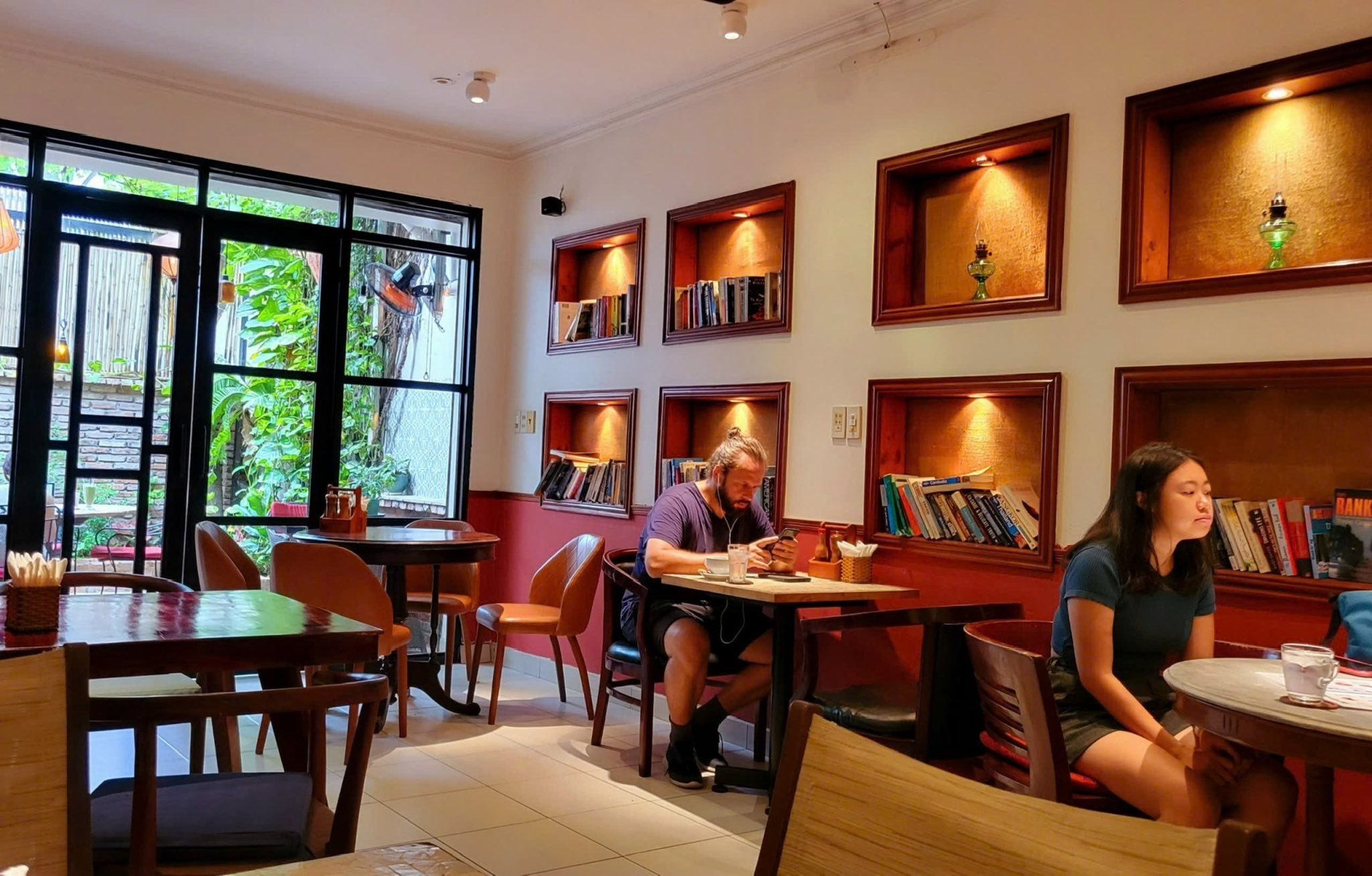 The image depicts an indoor setting of a cozy café or restaurant with red walls and wooden shelves displaying books. There are several patrons seated 