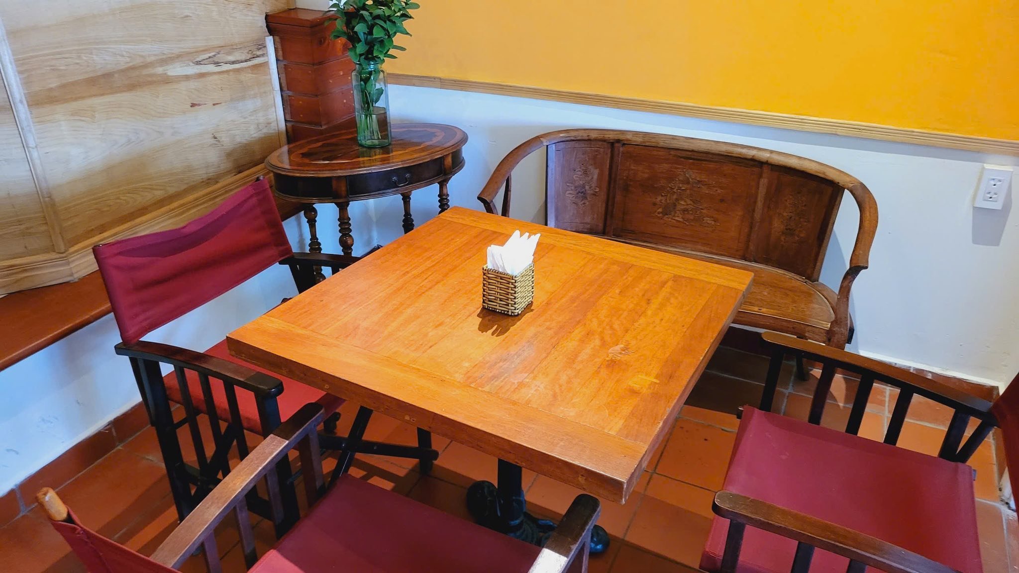 The image shows a cozy corner of an interior space featuring wooden furniture including chairs and tables with red cushions. A side table holds a vase
