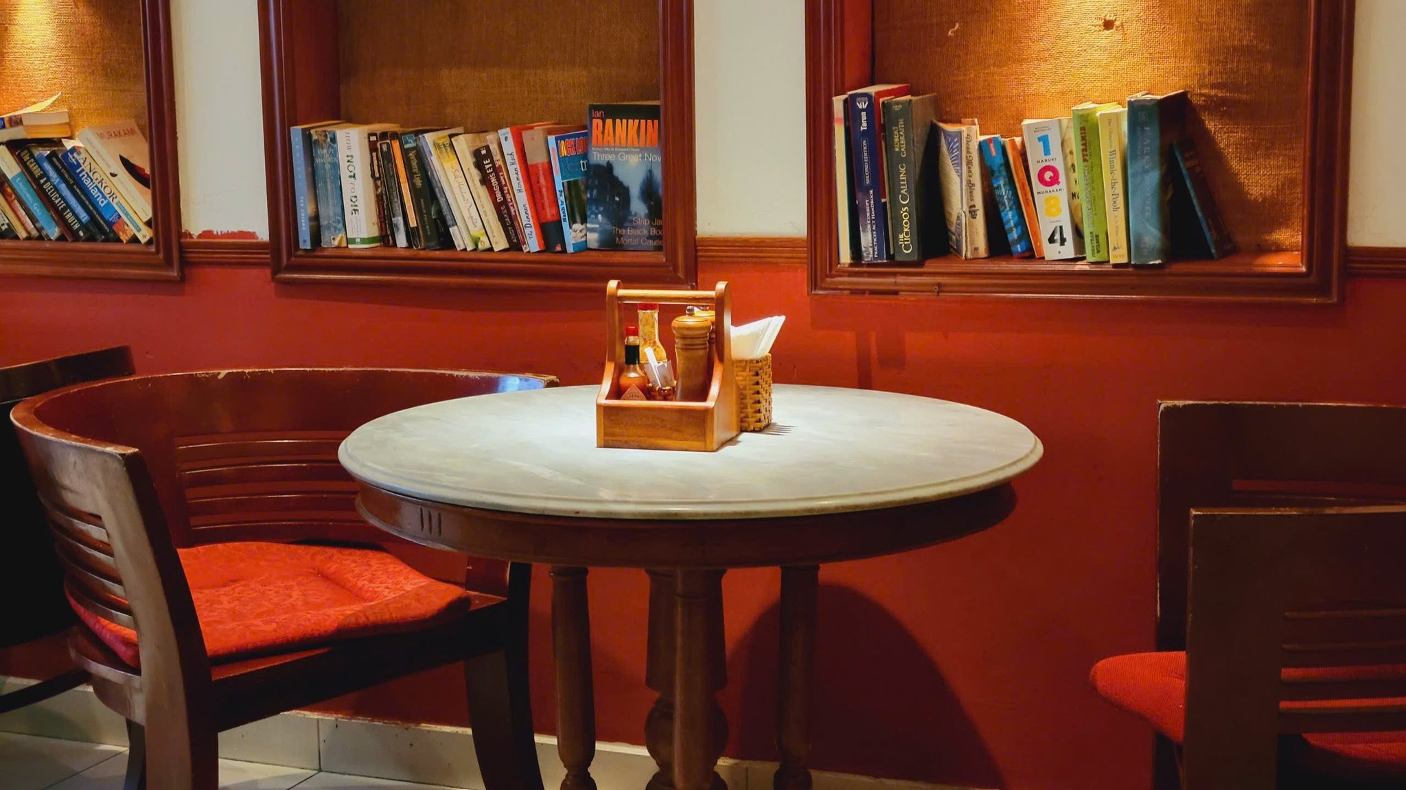 The image shows a cozy corner of an interior space featuring wooden chairs and tables against red walls. A small round table is set with condiments in