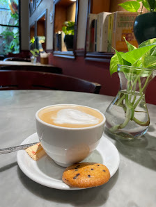 A cozy cafe setting featuring a table with a bowl of cappuccino and a cookie on the side, accompanied by a glass vase containing green plants in the b