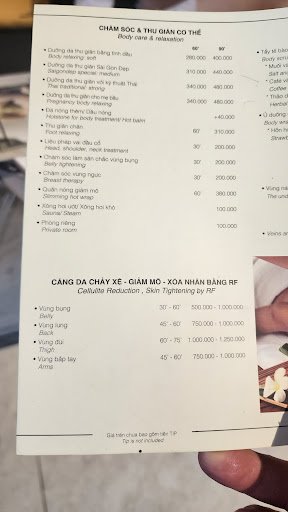 A person is holding a menu from 'Cham Doc & Thai Gian Co The' which lists various services offered by the establishment, including body care and relax