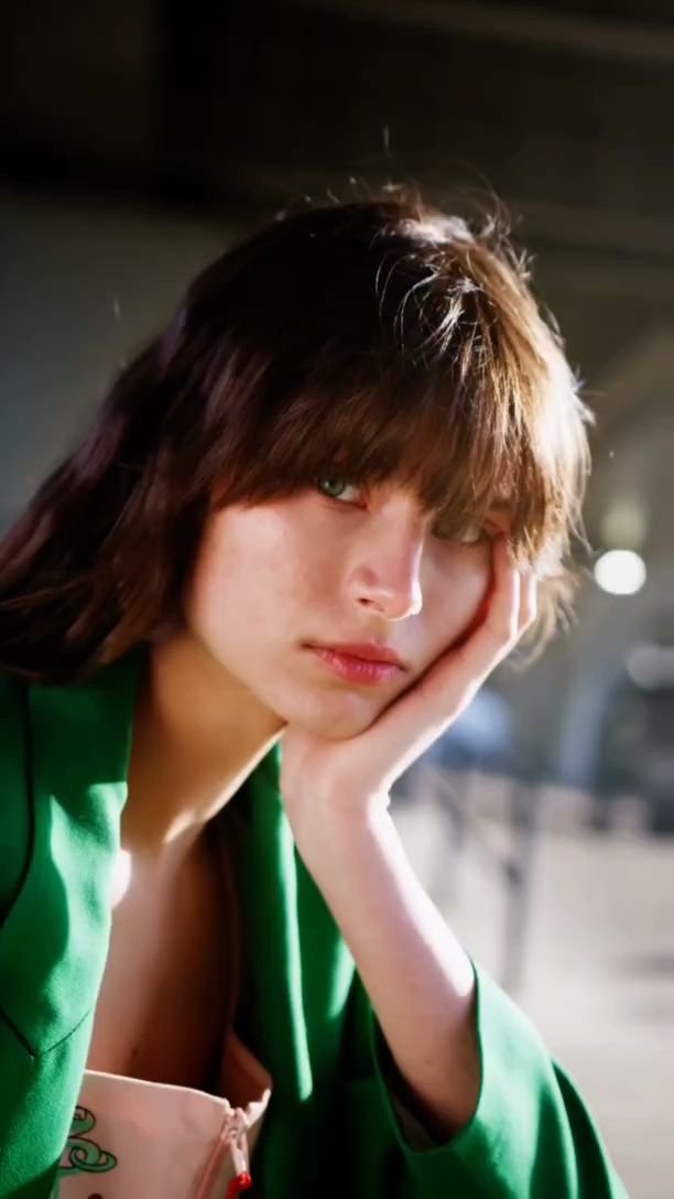 A person is seen in a dimly lit environment wearing a green garment and holding their head with one hand.