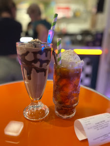 Two beverages are displayed on a bright orange surface in what appears to be an indoor setting with blurred background elements suggesting other patro