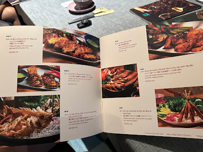 An open menu from a venue in Thao Dien, Saigon, displaying multiple food items with high-quality photographs and descriptive text. The menu features v