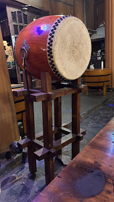 A traditional drum is displayed on a wooden stand in what appears to be an indoor setting with various items and furniture visible in the background.