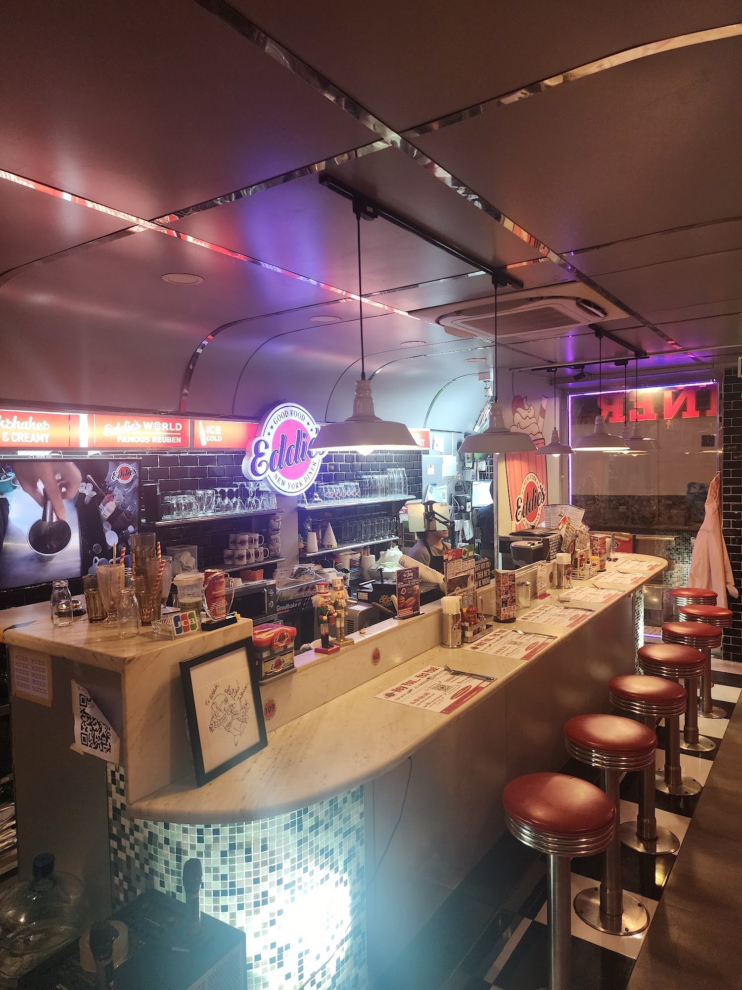 The image shows an interior view of a retro-themed diner-style venue named 'Edelweiss'. The decor includes neon signs, vintage stools, and tiled walls