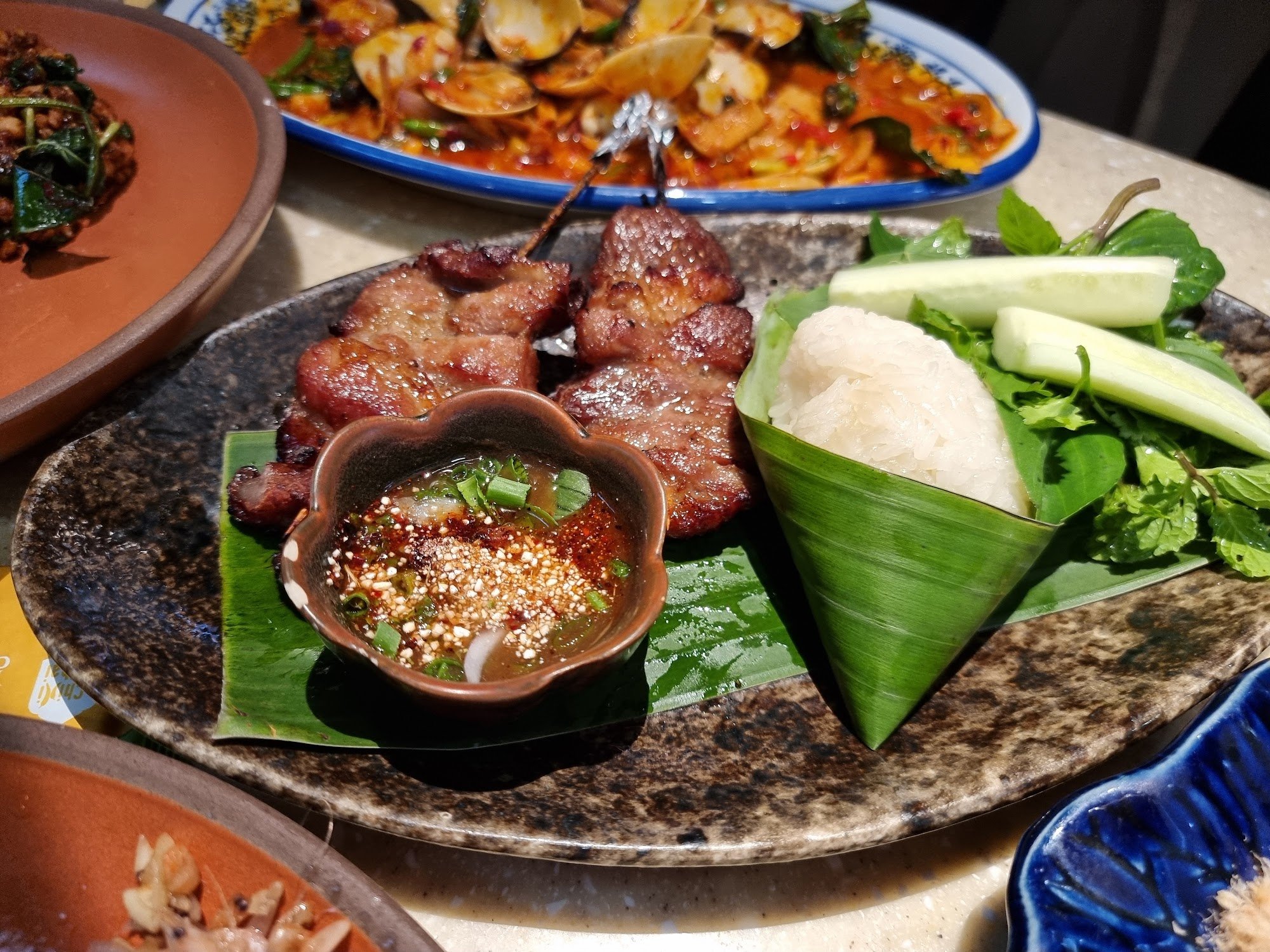 A close-up of a plate containing grilled meat skewers served on green banana leaves with a side of white rice and fresh vegetables, accompanied by a s
