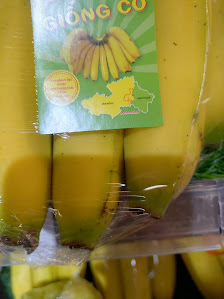 A bunch of yellow bananas in a plastic package labeled 'GIONG CO' is displayed prominently against the backdrop of what appears to be an interior sett