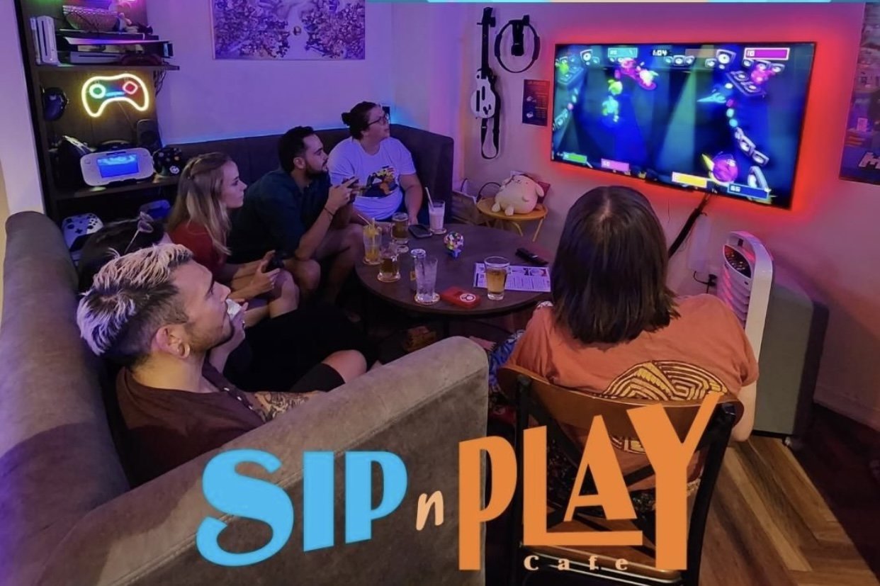 A group of people are gathered in a cozy room engaging in video gaming and socializing around a table with drinks. The ambiance is casual, illuminated