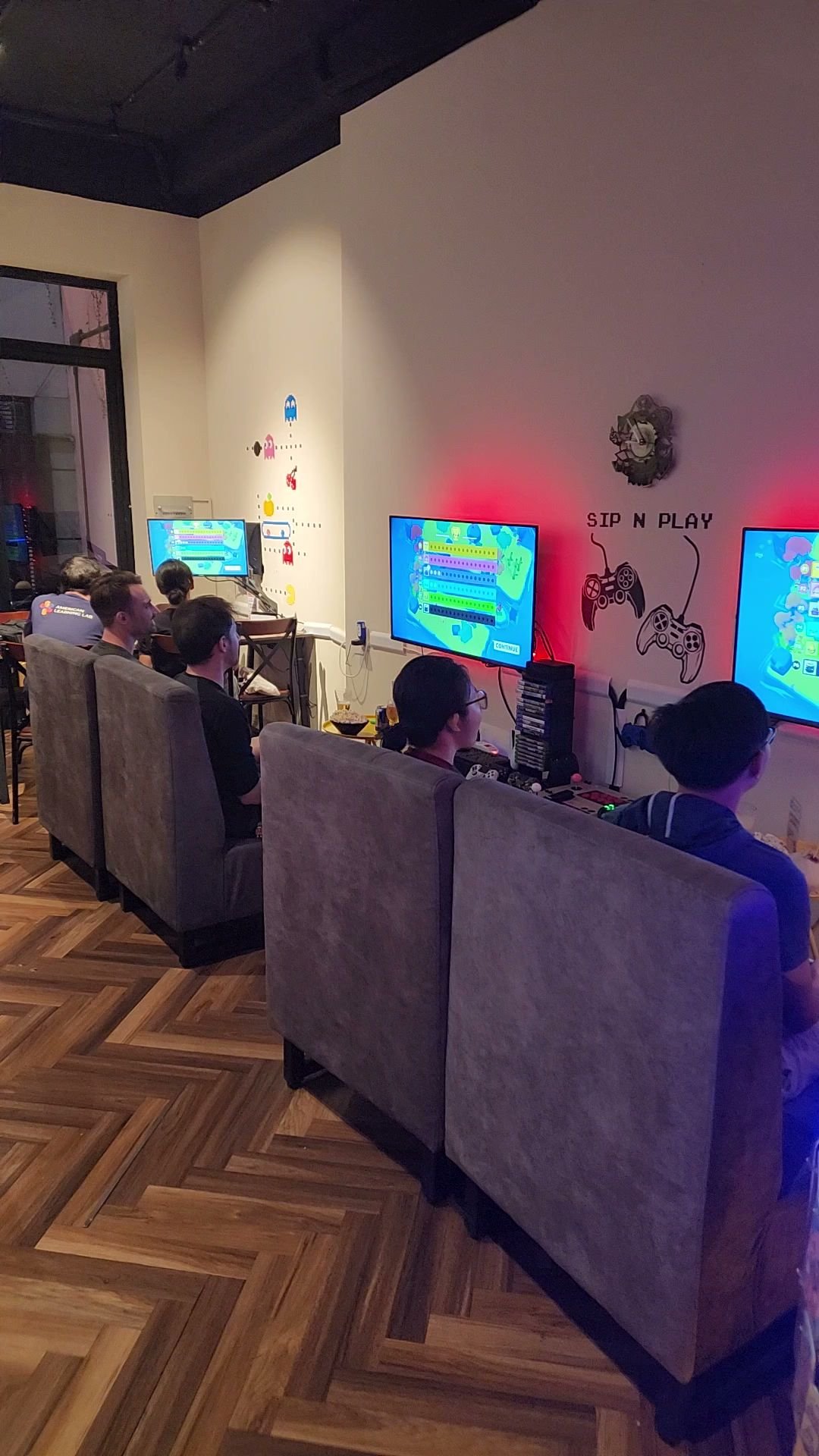 A group of people are seated on grey armchairs facing a row of monitors displaying colorful video game graphics in an indoor gaming area with wooden f
