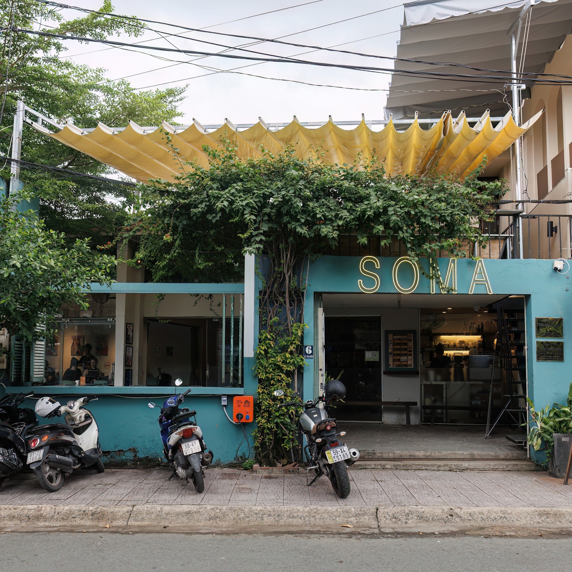 The image shows an exterior view of a cafe named 'SOMA' located in Thao Dien, Saigon. The establishment has a vibrant blue facade with greenery climbi
