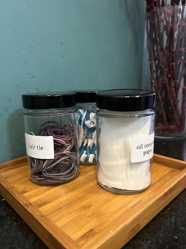 The image shows a wooden tray holding three glass jars labeled 'hair tie', 'oil remover pads', and another jar with blue items that could be cotton sw