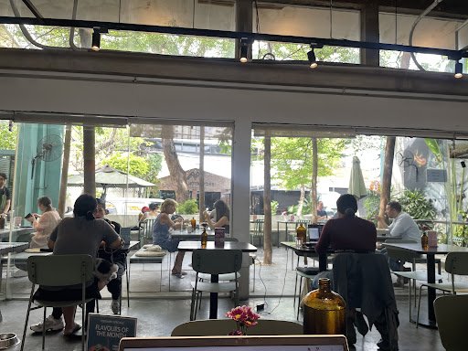 The image depicts an interior view of a modern cafe or restaurant in Thao Dien, Saigon. Patrons are seated at tables with various beverages and food i