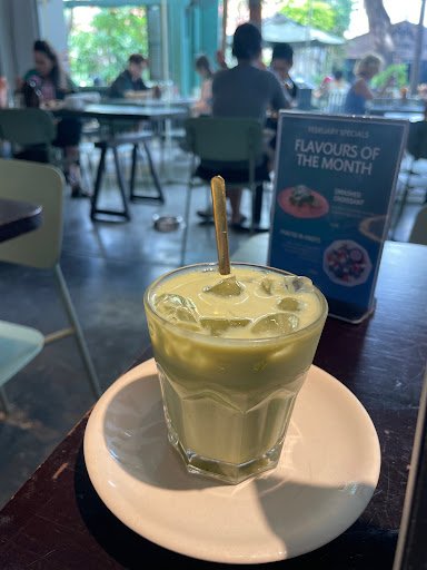 A glass of green tea beverage topped with ice cubes and a straw is placed on a white saucer in the foreground. In the background, there's an interior 