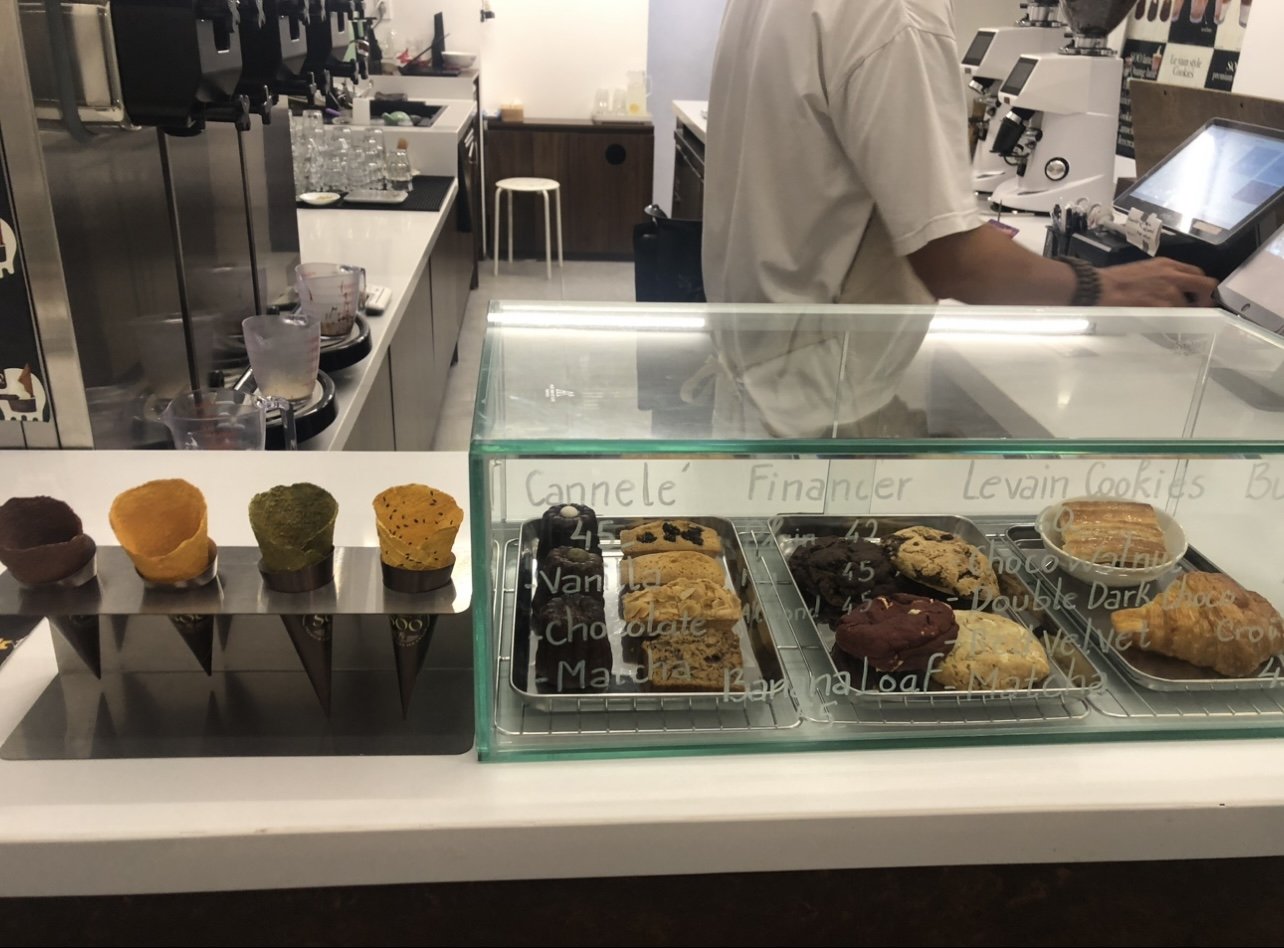 The image shows an interior view of a coffee shop or bakery counter area where various baked goods and drinks are displayed for sale.