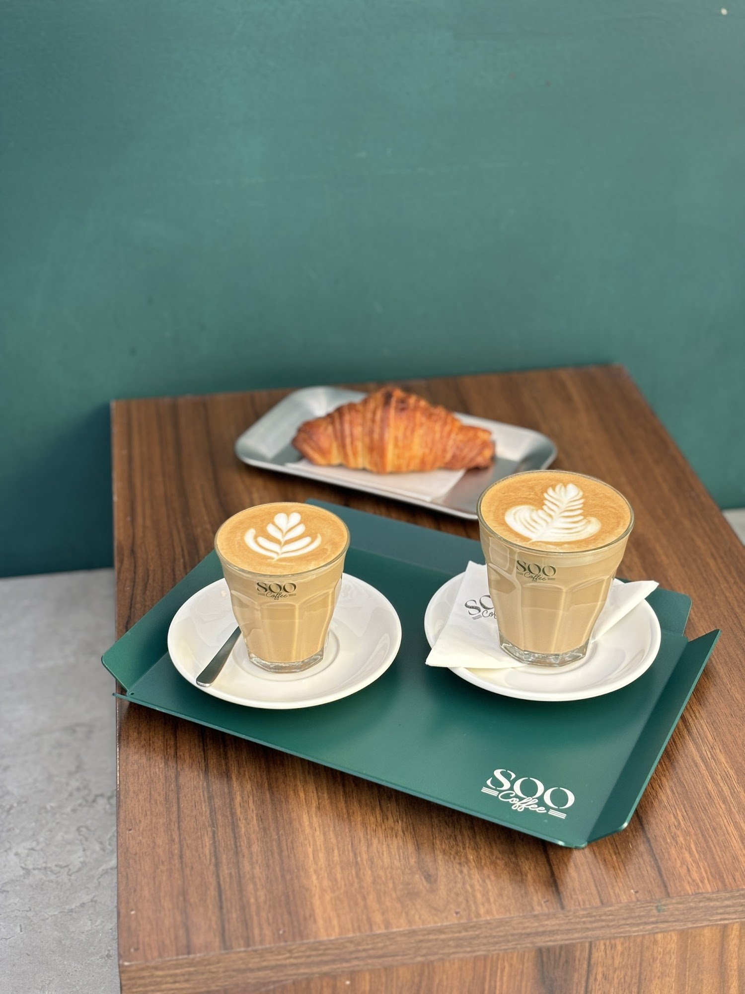 The image shows a neatly arranged coffee and croissant set on a wooden table against a green wall in what appears to be an interior setting of Thao Di