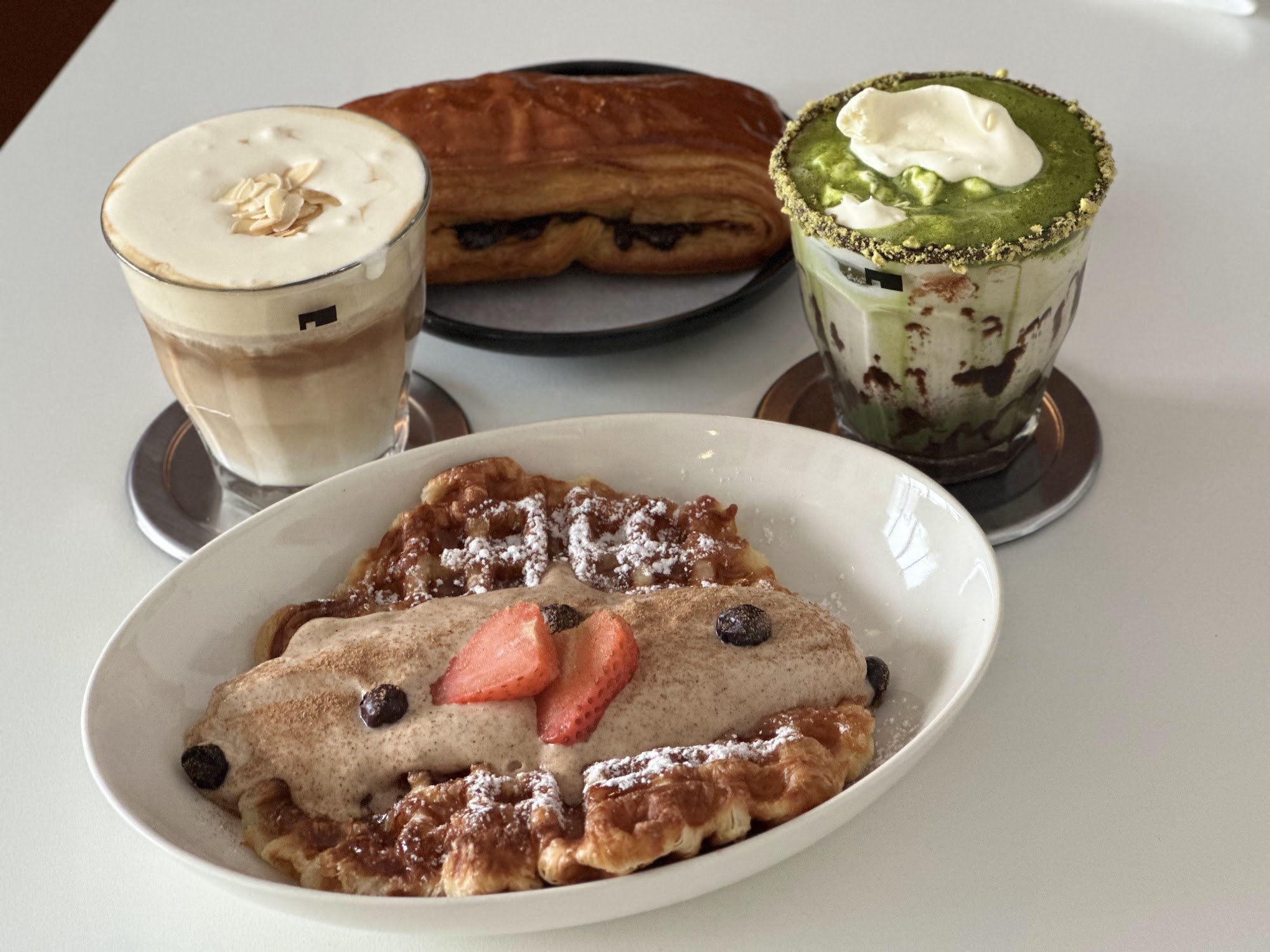 A plate of crepes or a similar pastry topped with powdered sugar, strawberries, and chocolate chips. Accompanied by two beverages: one appears to be an iced coffee drink garnished with whipped cream and nuts on top, while the other is a green tea latte with a dollop of whipped cream.