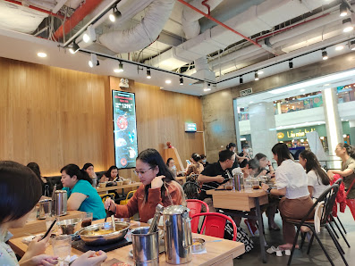 A bustling restaurant interior in Thao Dien, Saigon, with multiple patrons seated at tables. Features include wooden panel walls, red chairs, metal ta