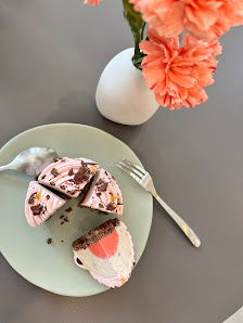 A slice of pink and white layered cake on a green plate accompanied by a fork is placed next to it with a vase containing orange flowers in the backgr